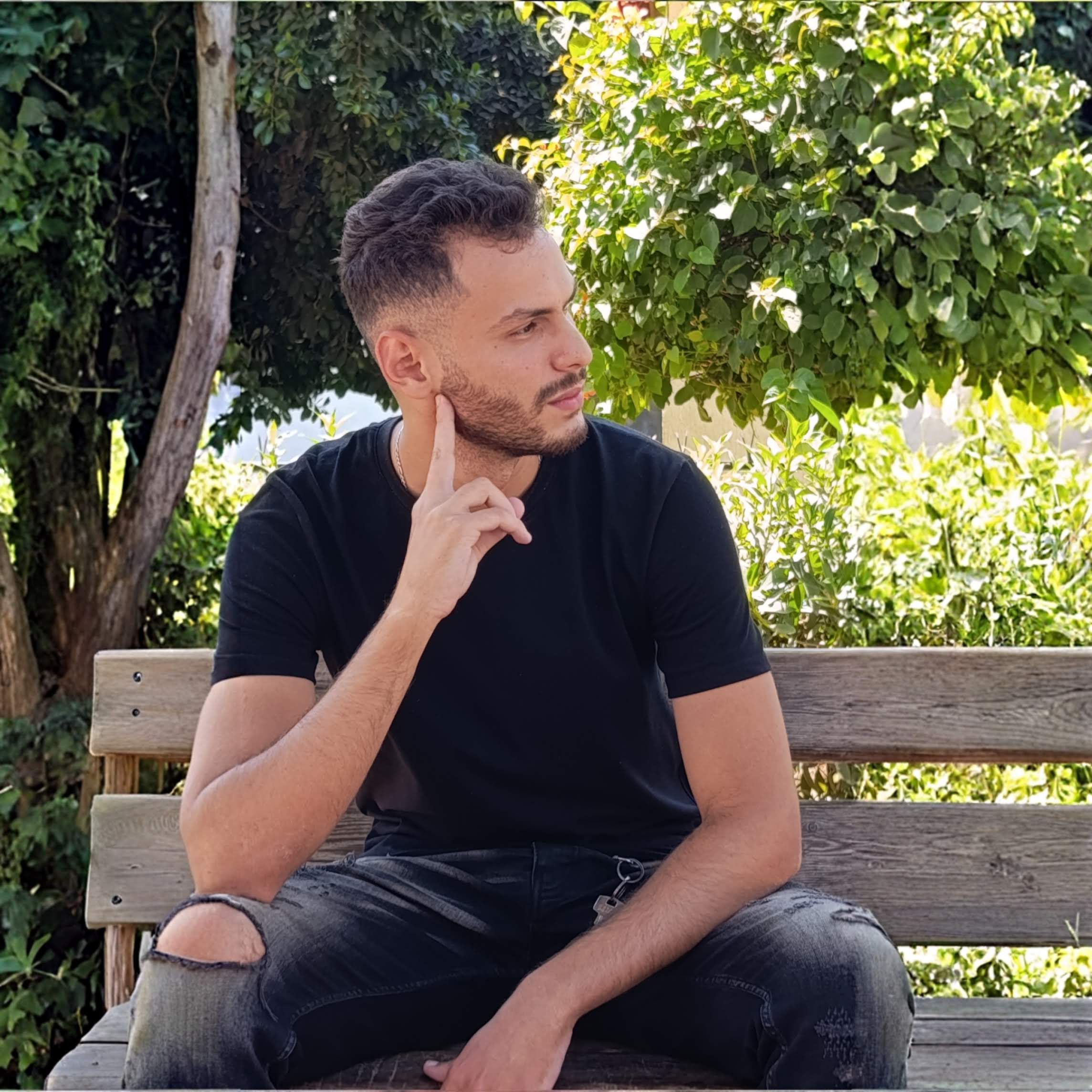 Side image of Adel Amrane with a black short sleeve t-shirt sitting in a bench in a park.